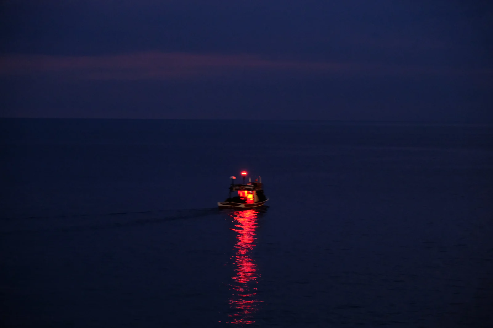 Gece denizinde tek başına duran, kırmızı ışığı suya uzun bir iz bırakan küçük bir tekne.