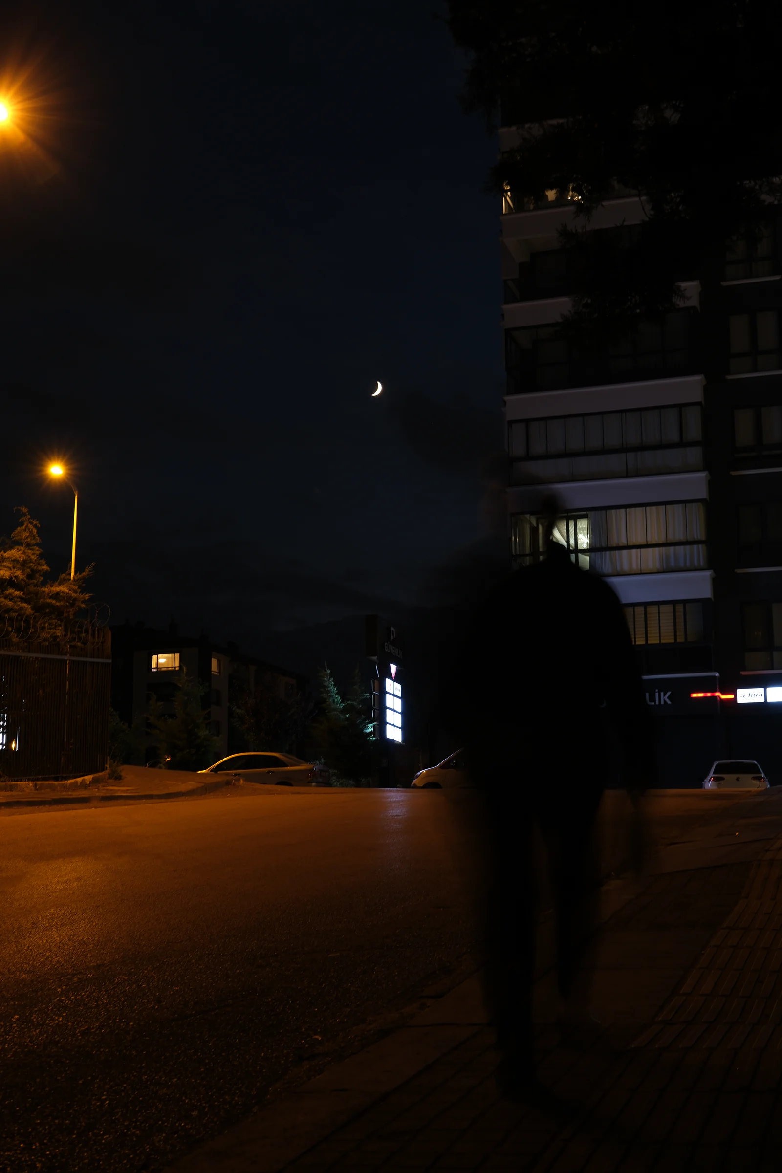Ay ve sokak lambasının altında, boş görünen bir sokaktan geçen hareketli bir figür.